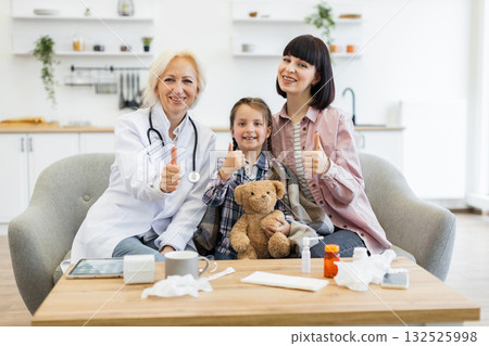 A doctor, a mother, and a young girl are sitting on a couch, smiling and giving thumbs up after a successful medical checkup in a home setting. 132525998