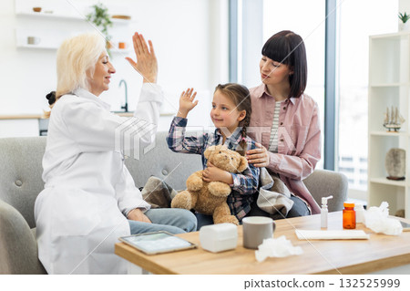 A doctor gives a high five to a young girl, who is sitting with her mother on a sofa, possibly after a checkup. The girl holds a teddy bear. A doctor gives a high five to a young girl, who is sitting with her mother on a sofa, possibly after a checkup. The girl holds a teddy bear. 132525999
