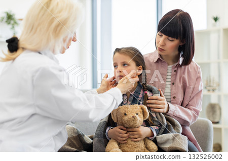 A doctor checks a young girl's throat while her mother comforts her. The girl is wrapped in a blanket, holding a teddy bear. A doctor checks a young girl's throat while her mother comforts her. The girl is wrapped in a blanket, holding a teddy bear. 132526002