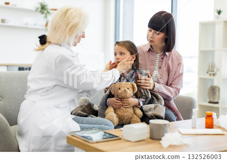 A doctor is checking a young girl's throat while her mother comforts her. The child is wrapped in a blanket and holding a teddy bear. A doctor is checking a young girl's throat while her mother comforts her. The child is wrapped in a blanket and holding a teddy bear. 132526003