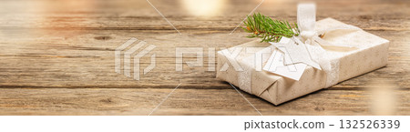 Elegant Christmas gift wrapped in beige paper with silver ribbon, fir branch, glitter star tag on wooden table with blurred warm lights copy space cozy holiday composition 132526339