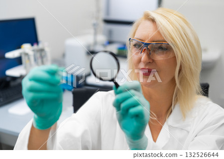 Middle aged female chemist in laboratory examining chemical solutions in test tubes with magnifying glass. Middle aged female chemist in laboratory examining chemical solutions in test tubes with magnifying glass. 132526644