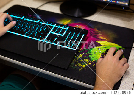Close up of hand using mouse on gaming mousepad with vibrant colors, illuminated keyboard with blue backlight. Modern gaming setup in home office desk 132528936