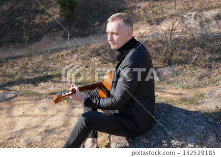 Blonde man playing string guitar outdoors in autumn forest. Concept of sound therapy, mental health and wellness rituals. Calmness tranquility audio-sensory practices. Aura farming energy 132529185