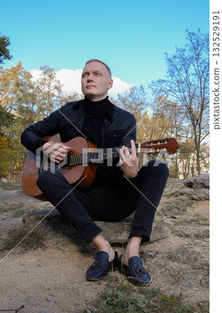 Beautiful blonde man playing acoustic guitar and singing relaxing enjoying outdoors on sunny autumn day. Artist learning and practicing playing musical instrument. Fall leaves autumnal 132529191