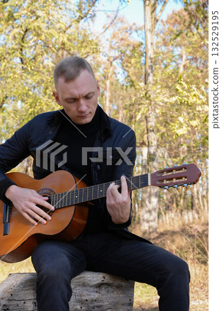 Portrait of smiling caucasian artist plays wooden acoustic guitar in autumnal park. Young songwriter plays string musical instrument hobby outside in nature fall time. Audio music healing Portrait of smiling caucasian artist plays wooden acoustic guitar in autumnal park. Young songwriter plays string musical instrument hobby outside in nature fall time. Audio music healing 132529195