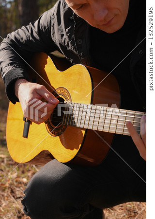 Beautiful blonde man playing acoustic guitar and singing relaxing enjoying outdoors on sunny autumn day. Artist learning and practicing playing musical instrument. Fall leaves autumnal 132529196