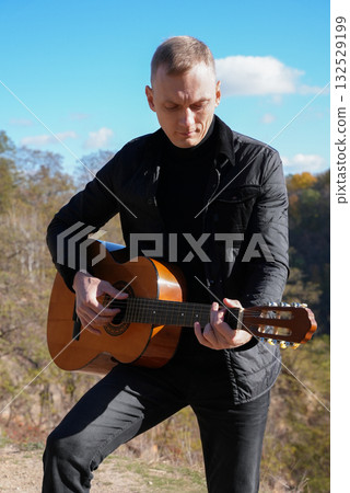 Beautiful blonde man playing acoustic guitar and singing relaxing enjoying outdoors on sunny autumn day. Artist learning and practicing playing musical instrument. Fall leaves autumnal 132529199