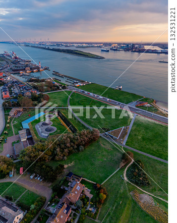 Aerial view of river port at sunset cargo ships, cranes, and industrial docks line both banks, with dense urban grid and green fields framing the waterway in a dynamic commercial landscape. 132531221