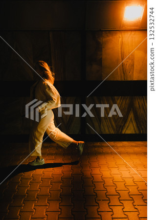 Woman in white suit is running in pedestrian underpass at night, illuminated by orange light, creating long shadow on ground. Woman in white tracksuit running through dimly lit pedestrian underpass 132532744