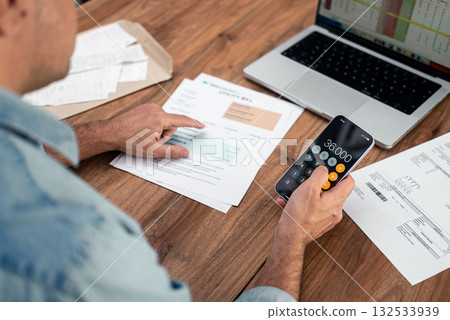 Man calculating utility bills using a smartphone calculator and reviewing documents on a wooden desk. Concept of household expenses, financial management, and budget planning.  132533939