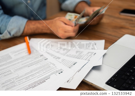 Man in denim shirt counting cash while examining tax forms on a wooden desk. Tax preparation and financial management concept, ideal for accounting and finance themes  132533942