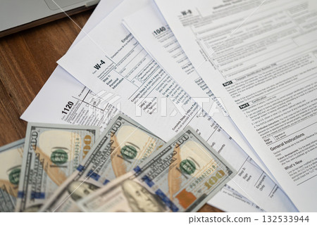 Tax forms and dollar bills spread out on a wooden desk near a laptop. Financial management, tax refund, and accounting concept.  132533944