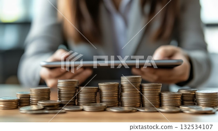 Businesswoman using digital tablet analyzing increasing stacks of coins, symbolizing investment growth, financial success, and wealth management 132534107