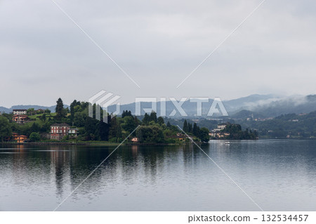 Calm morning view of Lake Orta with misty green hills and villas reflected in still water 132534457