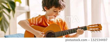 Happy child playing an acoustic guitar and singing joyfully at home, sitting comfortably on a sofa near a window, basking in warm natural light during a cheerful daytime 132534497