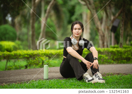 Young woman resting amidst lush greenery in a park, enjoying nature while wearing headphones, showcasing a relaxed lifestyle with fitness accessories 132541163