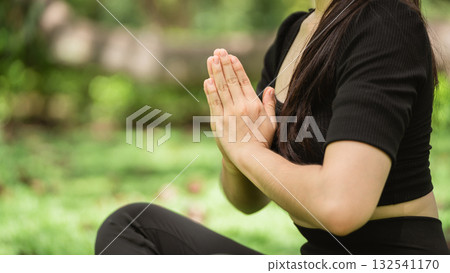Peaceful young woman practicing yoga in nature with hands in prayer pose, surrounded by greenery and soft sunlight filtering through leaves 132541170