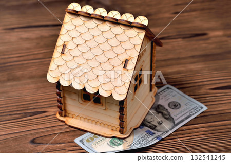 A small wooden house-piggy bank and a money bill of 100 dollars on a wooden brown background A small wooden house-piggy bank and a money bill of 100 dollars on a wooden brown background 132541245