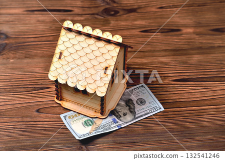 A small wooden house-piggy bank and a money bill of 100 dollars on a wooden brown background A small wooden house-piggy bank and a money bill of 100 dollars on a wooden brown background 132541246