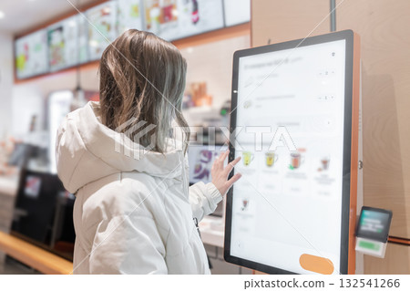 A woman uses a selfservice kiosk in a modern cafe, highlighting tech convenience in dining, A woman in a white jacket chooses drinks and orders food for dessert in a restaurant or pizzeria coffee shop 132541266