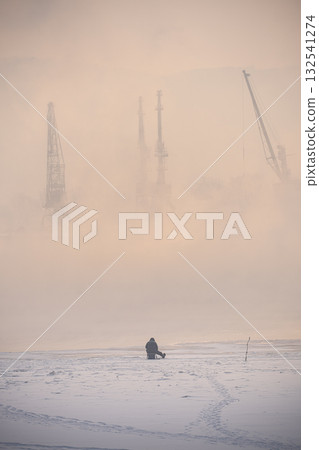 Winter fishing. A lone fisherman is fishing on the banks of the Yenisei River, against the background of port cranes visible through the fog in a pink mist.A misty winter landscape shows a solitary Winter fishing. A lone fisherman is fishing on the banks of the Yenisei River, against the background of port cranes visible through the fog in a pink mist.A misty winter landscape shows a solitary 132541274