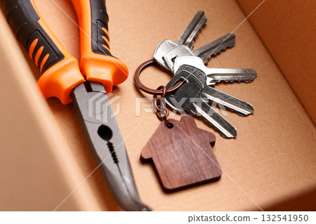 Curved nose pliers and bunch of door keys with wooden house keychain inside cardboard box. Symbol of home repair and DIY 132541950