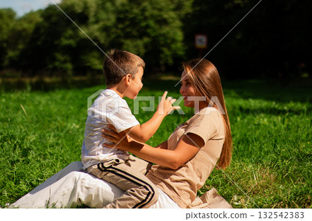 Candid portrait of happy mother and son sitting on green grass at public park on sunny summer day. Holidays with kids. Parenthood and motherhood topics. Candid portrait of happy mother and son sitting on green grass at public park on sunny summer day. Holidays with kids. Parenthood and motherhood topics. 132542383