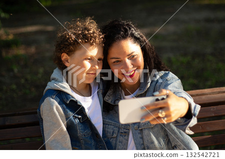 Young mother making selfie or video call with her son sitting on bench in park. Mom and boy calling dad. Concept of: connection, distance, technology 132542721