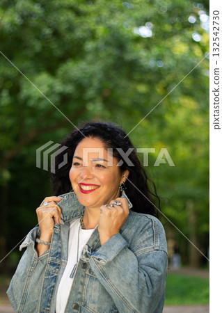 Portrait of confident curly female in nature. Close up portrait of dark haired woman with voluminous curls, smiling confidently against blurred green park background. Ideal for beauty, self love, and Portrait of confident curly female in nature. Close up portrait of dark haired woman with voluminous curls, smiling confidently against blurred green park background. Ideal for beauty, self love, and 132542730