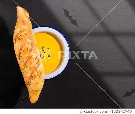 Halloween, food composition with home made pumpkin soup puree with fresh crunchy french baguette on a black background with bats shadows. Mystical, halloween, food card. Halloween, seasonal meal 132542740