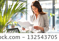 Businesswoman in bright office reviews documents with green plants and natural lighting in the background. Concept of eco-friendly workspace, focus, and professional approach 132543705