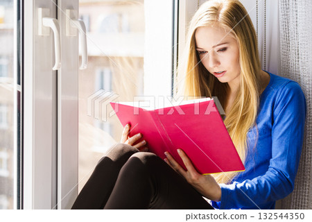 Woman sitting on window sill reading book at home 132544350