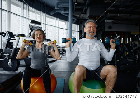 Mature couple sit on exercise balls in a gym with dumbbells Mature couple sit on exercise balls in a gym with dumbbells 132544778