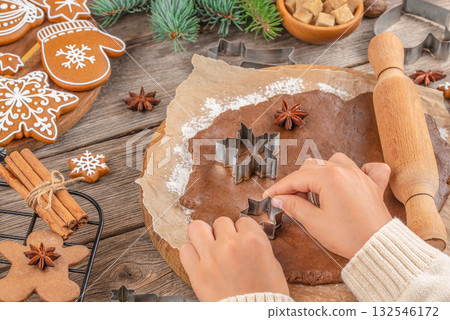 Hands cutting Christmas gingerbread cookies from dough with metal cutters on parchment paper, festive baking process with rolling pin, spices, decorated cookies, and fir branches, top view on table 132546172