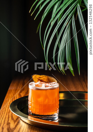 Vibrant orange cocktail with ice cubes and dried pear garnish served on a brass tray, illuminated by warm light and tropical leaves in the background 132546298
