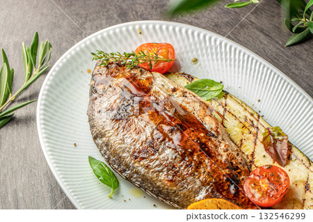 Grilled dorado fish served with zucchini, cherry tomatoes, and basil leaves, drizzled with olive oil and herbs on a white plate 132546299