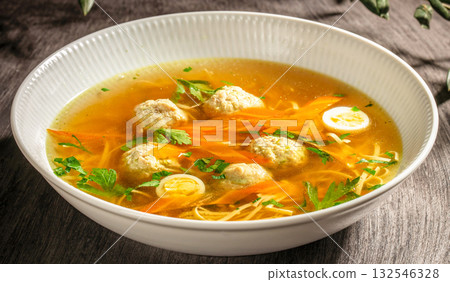 Homemade chicken broth with meatballs, noodles, carrots, and quail eggs served in a white bowl. 132546328