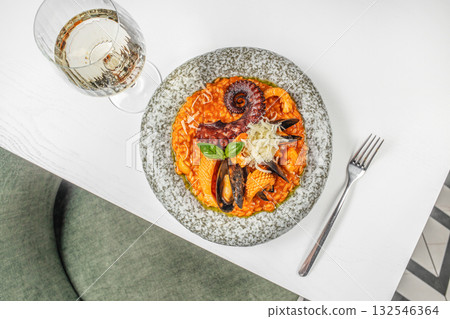 Flat lay of a gourmet seafood risotto with octopus tentacle, mussels, squid, tomato sauce, and grated parmesan cheese, served on a stone plate with a glass of white wine. Mediterranean food Flat lay of a gourmet seafood risotto with octopus tentacle, mussels, squid, tomato sauce, and grated parmesan cheese, served on a stone plate with a glass of white wine. Mediterranean food 132546364