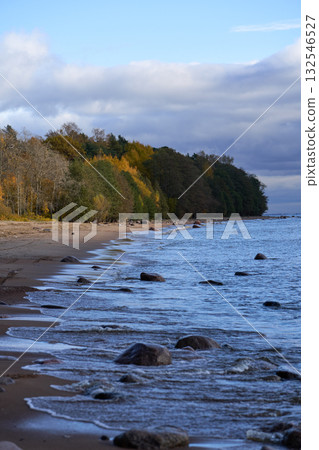 Waves hitting stones along autumn forest shoreline on calm sea. Waves hitting stones along autumn forest shoreline on calm sea. 132546527
