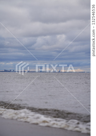 Distant city skyline and modern tower viewed across calm sea under cloudy sky. 132546536