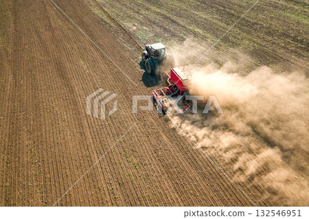 Tractor cultivating ground and seeding a dry field. Farmer preparing land with seedbed cultivator as part of pre seeding activities in early spring season of agricultural works at farmlands. Tractor cultivating ground and seeding a dry field. Farmer preparing land with seedbed cultivator as part of pre seeding activities in early spring season of agricultural works at farmlands. 132546951