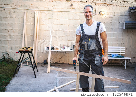 Mature male carpenter using electric screwdriver on a wooden plank outdoors 132546971