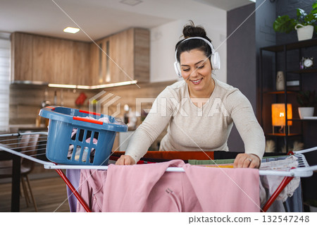 Woman listening to music while hanging the laundry on drying rack at home Woman listening to music while hanging the laundry on drying rack at home 132547248