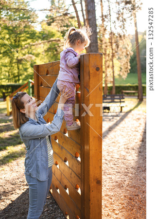 Smiling mom helps her cute daughter climb the playground Smiling mom helps her cute daughter climb the playground 132547285