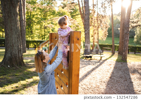 Smiling mom helps her cute daughter climb the playground Smiling mom helps her cute daughter climb the playground 132547286