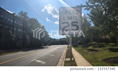 A 25 mph speed limit sign on a road between a park and an apartment building in the United States on a sunny day, American traffic rules and speed regulations, road signs on American urban highways A 25 mph speed limit sign on a road between a park and an apartment building in the United States on a sunny day, American traffic rules and speed regulations, road signs on American urban highways 132547386