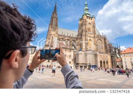 Prague, Czech Republic, August 8, 2023. View of St. Vitus Cathedral with the churchyard crowded with visitors. A young Caucasian man takes photos with his cell phone. A lifestyle using technology. Prague, Czech Republic, August 8, 2023. View of St. Vitus Cathedral with the churchyard crowded with visitors. A young Caucasian man takes photos with his cell phone. A lifestyle using technology. 132547416