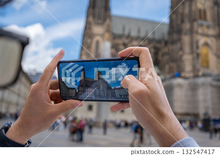 Prague, Czech Republic, August 8, 2023. View of St. Vitus Cathedral with the churchyard crowded with visitors. A young Caucasian man takes photos with his cell phone. A lifestyle using technology. Prague, Czech Republic, August 8, 2023. View of St. Vitus Cathedral with the churchyard crowded with visitors. A young Caucasian man takes photos with his cell phone. A lifestyle using technology. 132547417