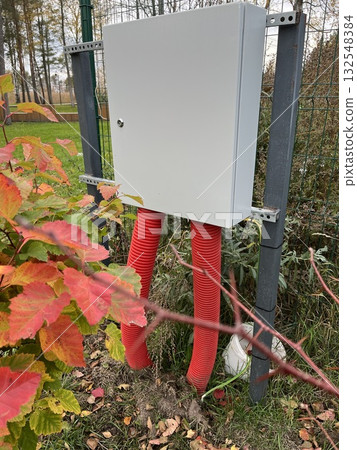 Electrical utility box mounted on metal supports with bright red conduits extending from the base, surrounded by autumn foliage and greenery, showcasing outdoor infrastructure 132548384